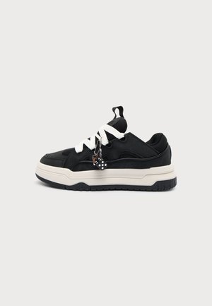 Zapatilla negra con suela blanca gruesa, cordones blancos y pequeños adornos de dados en blanco y negro colgando de los cordones sobre un fondo liso.