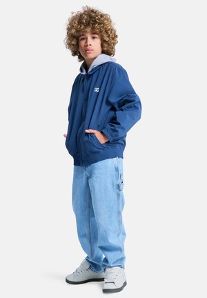 Marineblauw jack met een grijze capuchon, voorzien van een logo op de borst, gecombineerd met lichtblauwe wijde jeans en grijze hoge sneakers.