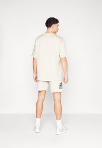 Ljust beige T-shirt med korta ärmar, matchad med shorts som har en svart grafiktryck på vänster ben. Slät bomullsmaterial.