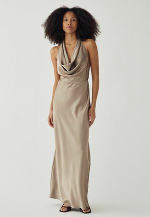 Kobieta z naturalnie kręconymi włosami, ubrana w długą beżową sukienkę na szyję typu halter oraz czarne sandały z odkrytymi palcami, stojąca na tle jednolitego jasnego tła.