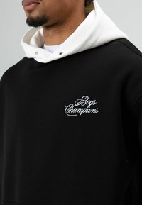 Sweat à capuche en coton noir avec une capuche blanche et un texte brodé "Boys Champions" en cursive blanche. Gros plan montrant la texture douce et les détails du design.