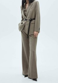 Jasnobrązowy kostium w kratkę z dopasowaną marynarką z paskiem i spodniami o szerokich nogawkach. Gładka tekstura z ostro zakończonymi czarnymi butami.