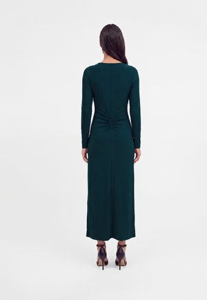 Femme portant une robe midi à manches longues vert foncé avec un détail froncé au bas du dos et des sandales à talons noires, debout de dos.
