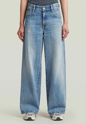 Persoon die lichtblauwe wijdvallende jeans en grijze sneakers draagt, staand tegen een effen achtergrond met ontspannen handen langs de zij.