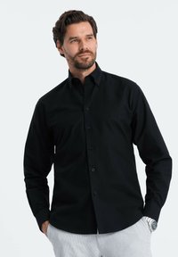 Camicia nera con bottoni realizzata in tessuto morbido, con maniche lunghe, colletto e silhouette aderente. Design semplice senza fantasie.