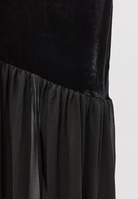 Czarna sukienka z welwetowym gorsetem i zwiewną, plisowaną spódnicą z przezroczystego materiału. Tekstura przechodzi od gładkiej do miękkiej.