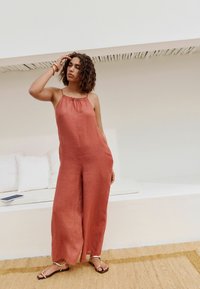 Jumpsuit od lanenog materijala boje rđe sa skupljenim izrezom, širokim nogavicama i podesivim trakama. Uparen sa ravnim sandalama sa zlatnim detaljima.