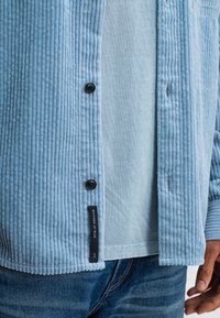 Light blue corduroy shirt with vertical stripes, black buttons, and a tag at the bottom reading "Butcher of Blue." Underlayer is light blue.