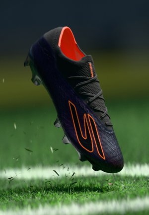 Scarpa da calcio nera con interno e logo arancione, upper testurizzato e suola a tacchetti moldati, catturata in aria sopra un prato verde.