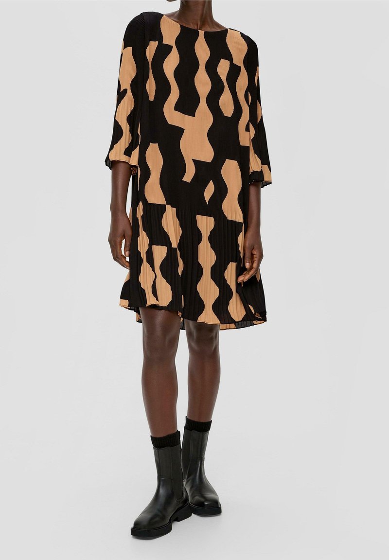 Femme portant une robe ample mi-longue avec des motifs abstraits noirs et beiges, associée à des bottines noires et des chaussettes noires.