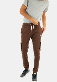 Pantalones de chándal de algodón marrón con bolsillos laterales, cintura con cordón y parche de logo. Combinados con una camiseta gris y zapatillas oscuras.