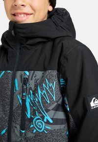 Schwarze Schneejacke mit geometrischem Druck in Grautönen und Blau, hochgeschlossenem Kragen und Reißverschlusstaschen.