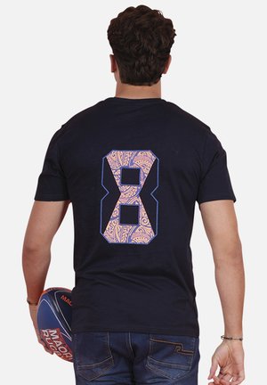 Homme vu de dos portant un t-shirt noir avec un grand numéro décoratif 8, tenant un ballon de rugby bleu et orange dans sa main gauche.