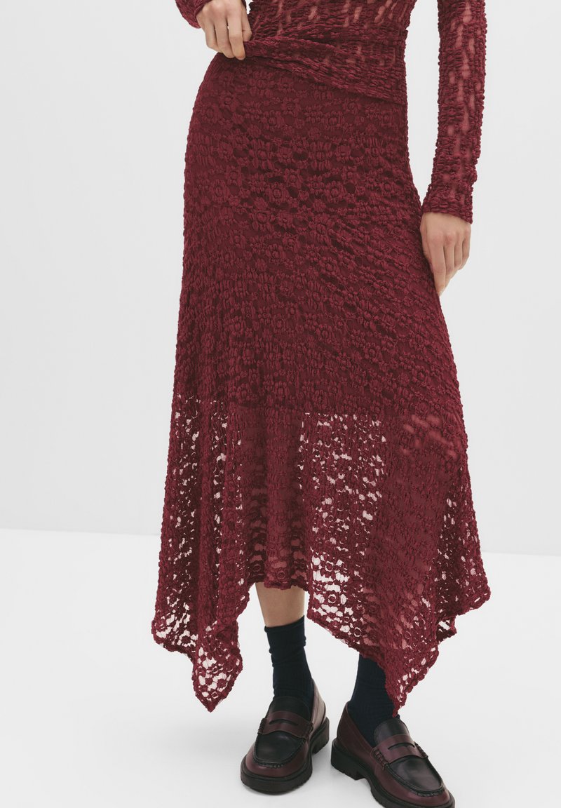 Burgunderfarbene Spitzenrock mit gewelltem Saum und strukturiertem Blumenmuster. Kombiniert mit schwarzen Mokassins und marineblauen Socken, mit einem eng anliegenden Bund.