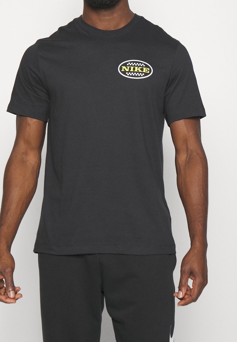 T-shirt noir à manches courtes avec petit logo ovale Nike sur le côté gauche de la poitrine, porté par une personne debout à la peau foncée et aux bras musclés.