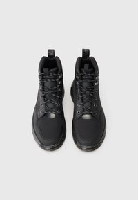 Ett par svarta högklackade sneakers med mönstrade snören och dragremmar på en vit bakgrund.