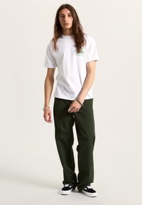 Vit bomull t-shirt med grön grafisk tryck, kombinerad med lösa mörkgröna byxor och svarta sneakers med vita detaljer.