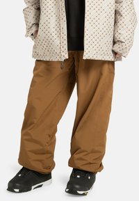 Bruine waterdichte broek met een losse fit, gecombineerd met een patroonjasje in beige met quiltstructuur en zwarte schoenen met witte accenten.