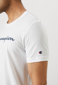 Witte katoenen t-shirt met een donkerblauw "Champion" logo op de voorkant en een klein rood en marineblauw logo op de mouw. Korte mouwen, ronde hals.