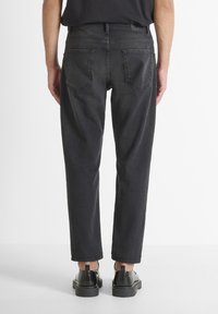 Jeans de mezclilla negra, corte relajado, cintura a media altura, con bolsillos traseros. Llevados con zapatos negros, mostrando un dobladillo ligeramente recortado. Textura mate.