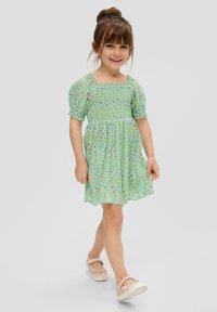 Hellgrünes kurzes Kleid mit schmalem Oberteil und Puffärmeln, ausgestattet mit einem blumigen Muster in Lila und Gelb. Kombiniert mit rosa Schuhen.