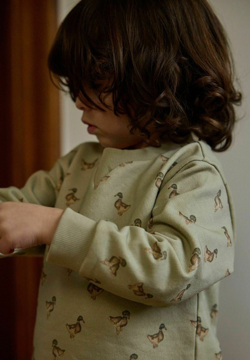 Jeune enfant aux cheveux bouclés et foncés portant un sweat-shirt vert clair avec de petits dessins de canards, regardant vers le bas et ajustant ses manches.