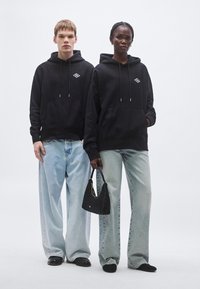 Čierne mikiny s kapucňou so stiahnutím a prednými vreckami, skombinované s bledomodrými širokými džínsami. Jeden model drží čiernu tkanú kabelku.
