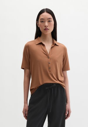 Jeune femme aux cheveux noirs raides portant une chemise marron à manches courtes avec col et un pantalon noir à cordon, sur un fond uni.