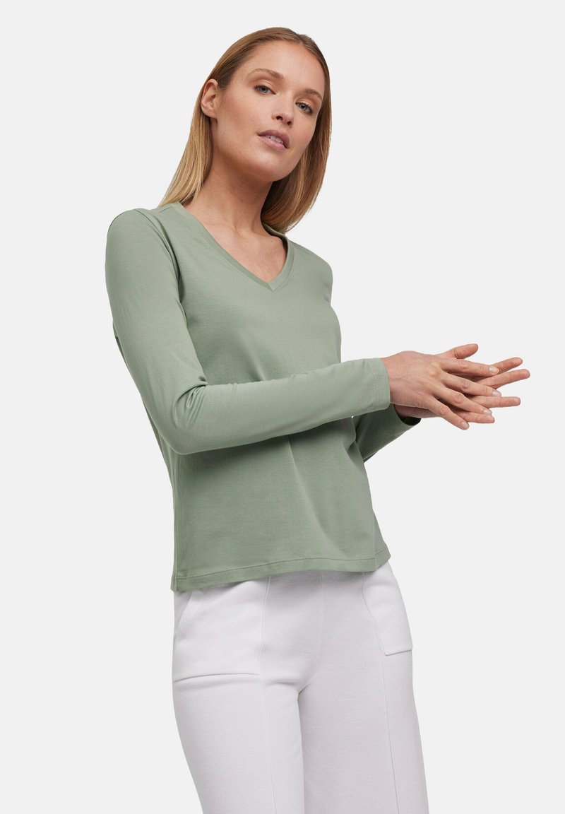 Langärmliges V-Ausschnitt-Shirt aus weichem grünem Stoff. Bietet eine lässige Passform und eine glatte Textur, kombiniert mit weißen Hosen für den Kontrast.