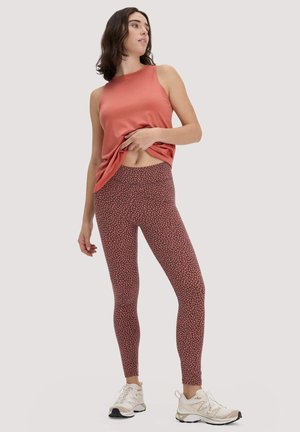 Vrouw draagt een mouwloze koraalkleurige top, legging met bloemenpatroon en witte sneakers, staand en haar top licht opheffend.