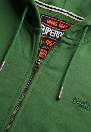 Groene rits hoodie met trekkoorden, metalen rits en trekkoorduiteinden met het label "Superdry," en een binnenlabel met de tekst "Superdry Goods Dept."