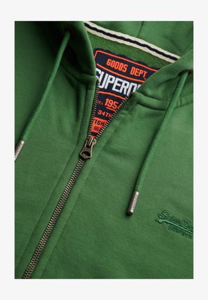 Groene rits hoodie met trekkoorden, metalen rits en trekkoorduiteinden met het label "Superdry," en een binnenlabel met de tekst "Superdry Goods Dept."