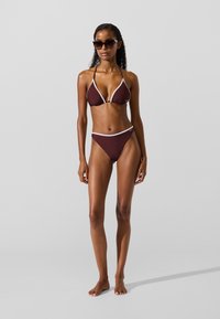 Bikiny sada v tmavohnedej farbe s bielym ozdobným lemovaním so zubovitým vzorom. Vrchný diel má trojuholníkové košíky; spodný diel je s nízkym pásom, oba vyrobené z hladkej, pružnej látky.