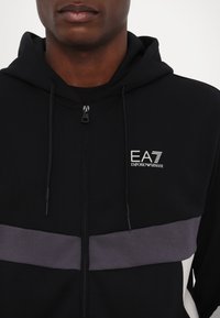 Black zip-up hoodie featuring a front logo, adjustable drawstring hood, and contrasting gray horizontal stripe across the chest.