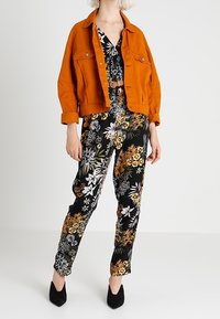 Giacca di jeans arancione sopra un tuta jumpsuit nera con stampa floreale; presenta vivaci motivi floreali in bianco, giallo e verde con gambe affusolate e tacchi neri.