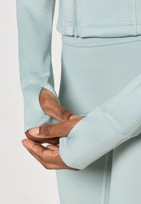 Σταυρωτό ενεργό μπουφάν ανοιχτού μπλε χρώματος με επεκταμένα μανίκια και τρύπες για τον αντίχειρα, κατασκευασμένο από ομαλό, ελαστικό ύφασμα με εμφανείς ραφές.