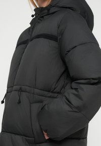 Black puffer jacket with a hood, featuring a front zipper, elastic drawstrings, and a quilted texture. Includes a contrasting black stripe.