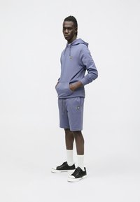 Lyse lilla hættetrøje med kængurulomme og broderet logo, kombineret med matchende shorts. Sorte sneakers og hvide sokker fuldender outfittet.