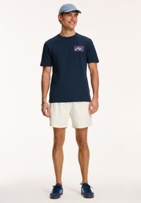 Tricou din bumbac bleumarin cu mâneci scurte și un accent grafic mic deasupra buzunarului stâng. Asortat cu pantaloni scurți texturati crem și adidași albaștri.