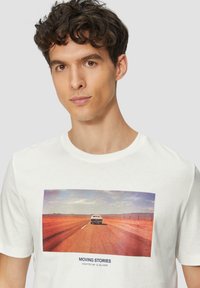 Witte katoenen t-shirt met een centrale afbeelding van een weg met een auto en een woestijnlandschap, met blauwe tekst onder de afbeelding.