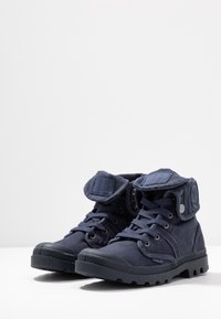 Botas de tobillo de lona azul marino con cordones en la parte delantera, cuello acolchado y suela de goma; cuenta con un parche de logo en el lado y una lengüeta plegable.