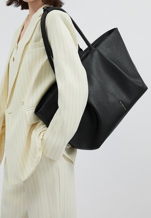 Frau trägt cremefarbenen Nadelstreifenanzug und große schwarze strukturierte Ledertasche auf der Schulter vor einfarbigem Hintergrund.