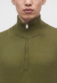 Gros plan d'une personne portant un pull zippé vert olive avec un col haut et le logo "HUGO" sur la poitrine.