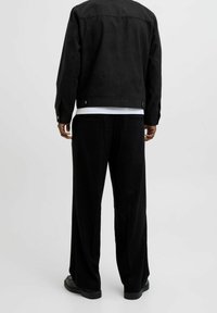 Czarna zamszowa kurtka o prostym kroju z zatrzaskami, zestawiona z szerokimi czarnymi sztruksowymi spodniami. Widok od tyłu ukazuje czysty design i fakturę.