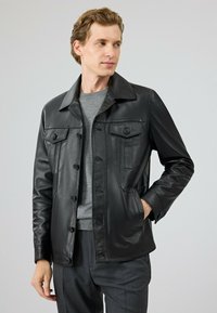 Zwarte leren jas met een knoopsluiting, twee borstzakken en een puntige kraag. Gedragen over een grijze trui met een zachte textuur.