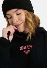 Felpa nera in pile con logo "ROXY" rosa e design a cuore. Cordino elastico al collo e tessuto con texture ben visibile.