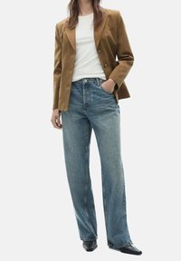 Femme portant un blazer marron sur une chemise blanche, un jean bleu taille haute et des chaussures noires pointues, debout avec une main dans la poche.