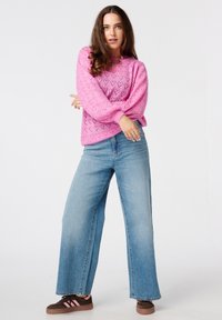 Roze gebreide sweater met hartpatronen, gecombineerd met lichtblauwe wijde jeans en bruine sneakers met roze accenten. Lange, losse mouwen.