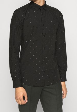 Mies, jolla on musta pikkutaskullinen paita, jossa on pieniä vaaleanpunaisia ja valkoisia pilkkukuvioita, ja tummanharmaat housut, seisomassa yksinkertaisen taustan edessä.