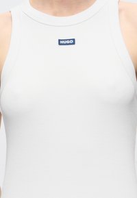 Gros plan d'une personne portant un haut blanc sans manches avec une petite étiquette bleue "HUGO" centrée près de l'encolure.